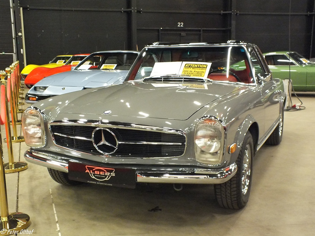 En vackert restaurerad silvergrå Mercedes-Benz klassisk roadster (troligen en 280SL Pagoda) utställd i en inomhusauktionslokal. Bilen har en 'ATTENTION'-informationsskylt på vindrutan, typiskt för auktionsmiljöer.